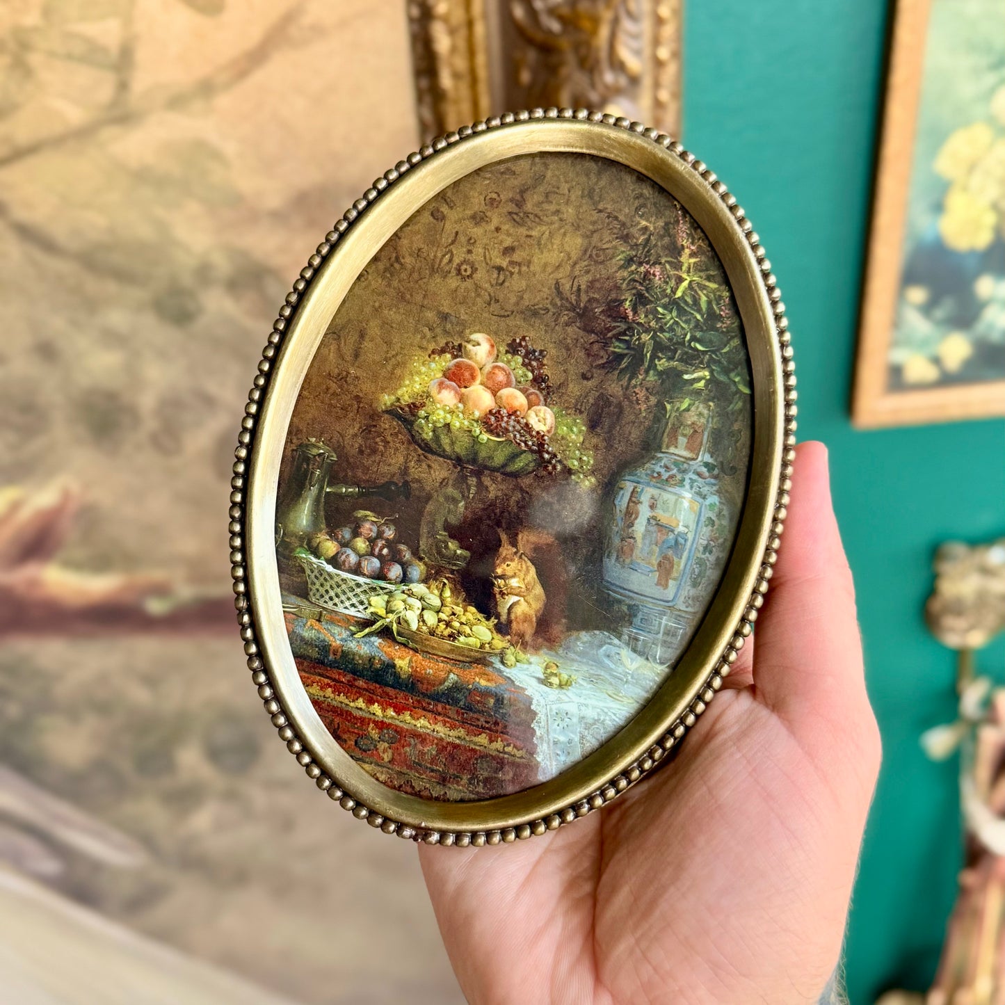 A small, antique-style framed artwork featuring a still life painting of a vase, fruit, and a squirrel. The painting is encased behind glass in an ornate, antiqued beaded brass frame.