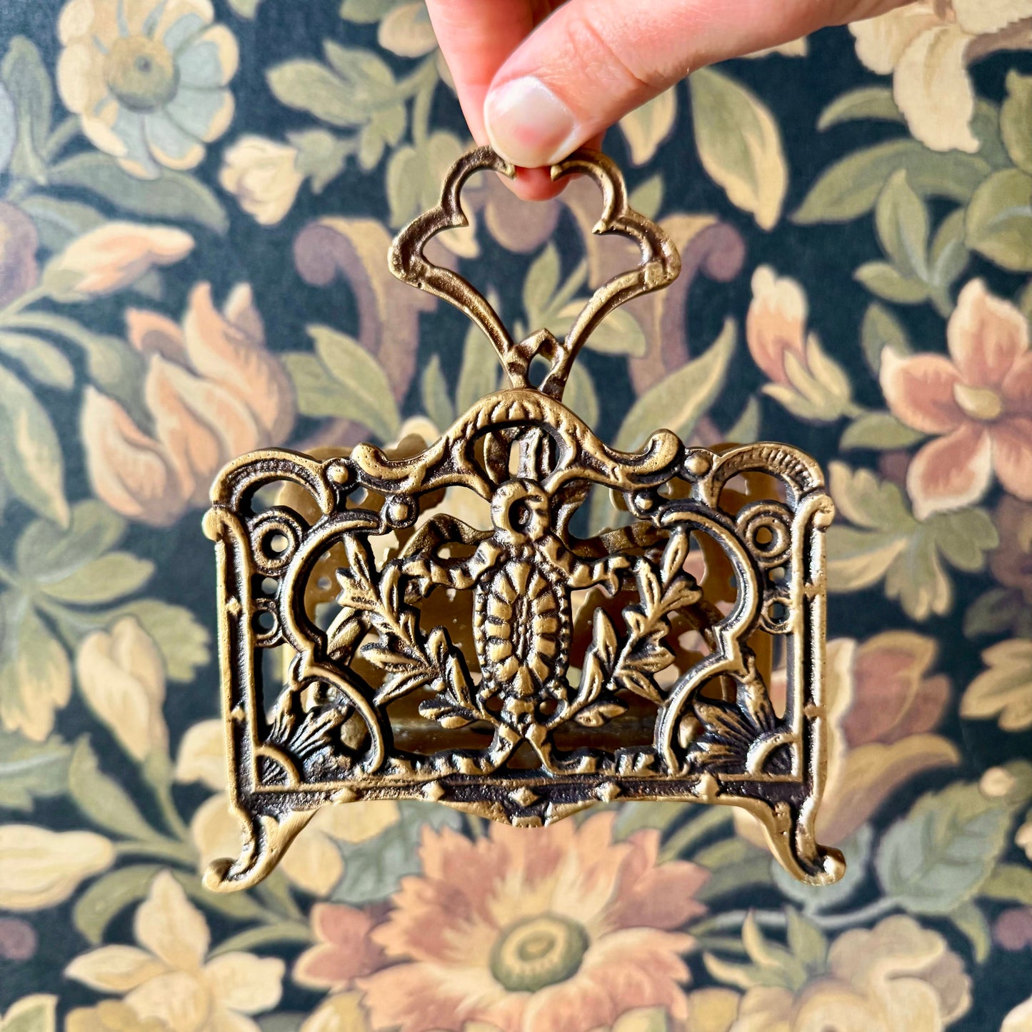 A detailed close-up of the Antique Brass Napkin and Letter Holder, showing its ornate curves and aged brass patina on a lace tablecloth.