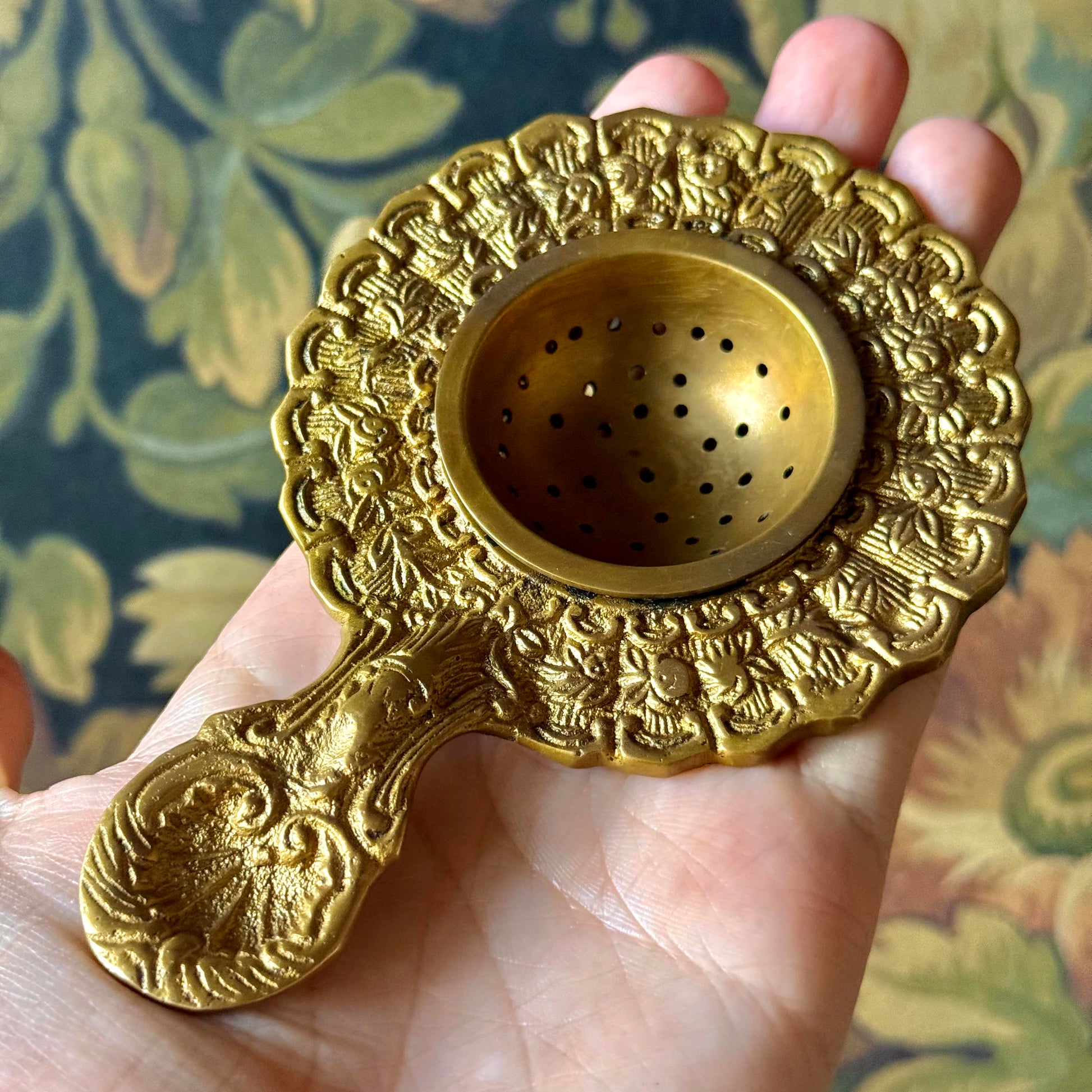 Antiqued brass tea strainer with intricate floral engravings and a scalloped edge, displayed on a neutral background. The vintage-style strainer is made of solid brass and measures approximately 5¼ inches long.