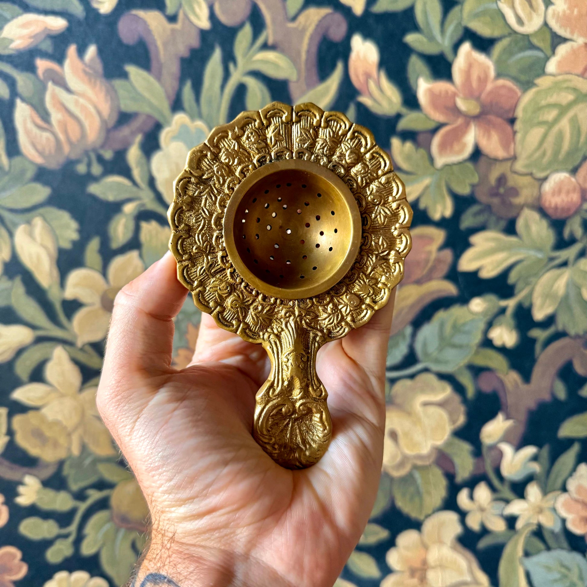 Antiqued brass tea strainer with intricate floral engravings and a scalloped edge, displayed on a neutral background. The vintage-style strainer is made of solid brass and measures approximately 5¼ inches long.