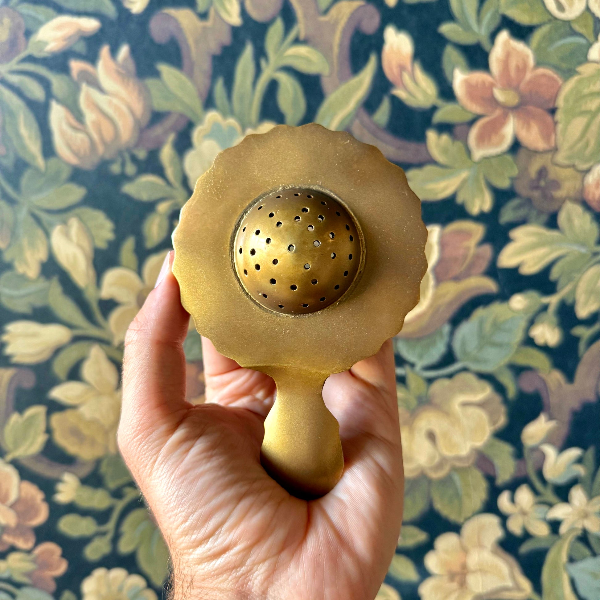 Antiqued brass tea strainer with intricate floral engravings and a scalloped edge, displayed on a neutral background. The vintage-style strainer is made of solid brass and measures approximately 5¼ inches long.