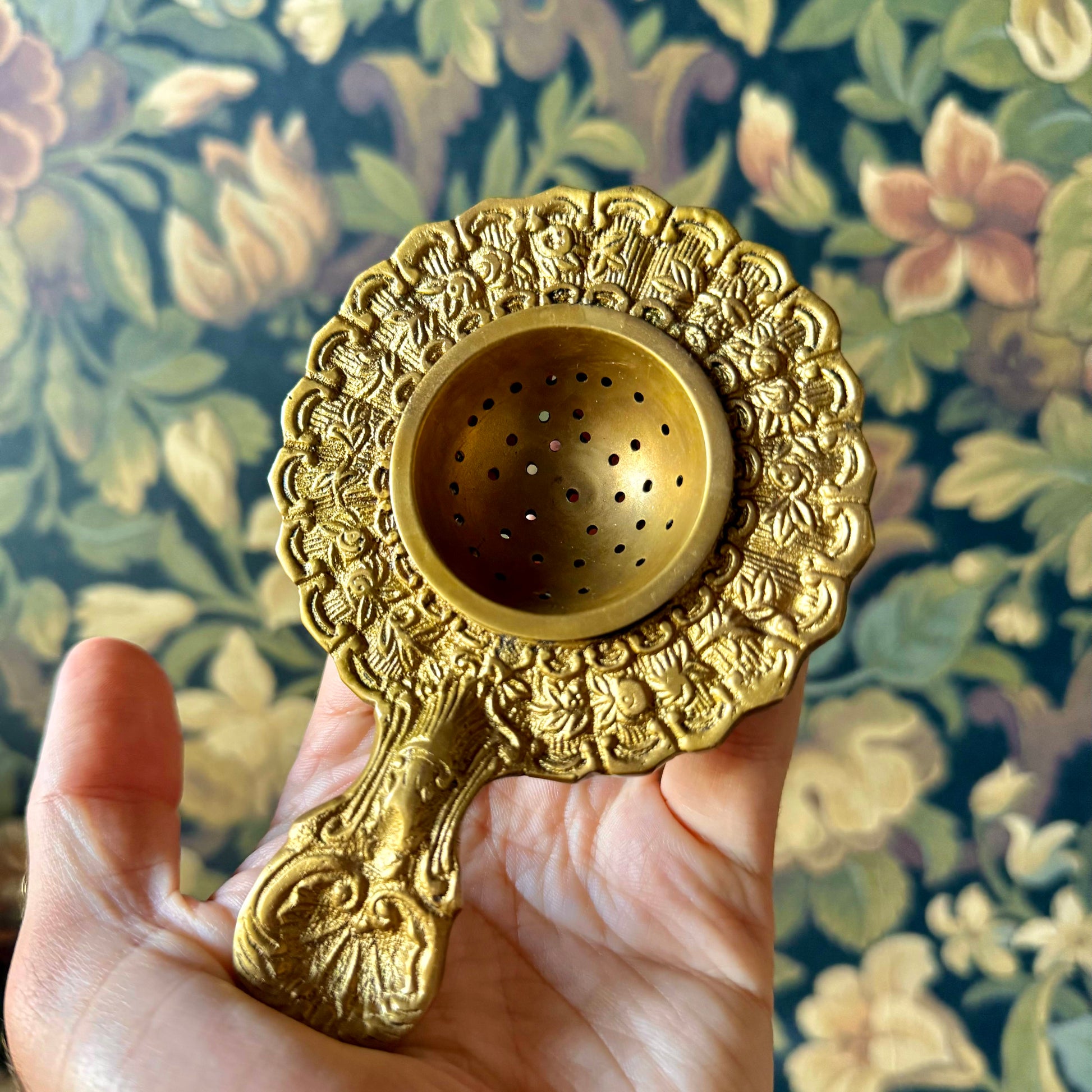 Antiqued brass tea strainer with intricate floral engravings and a scalloped edge, displayed on a neutral background. The vintage-style strainer is made of solid brass and measures approximately 5¼ inches long.