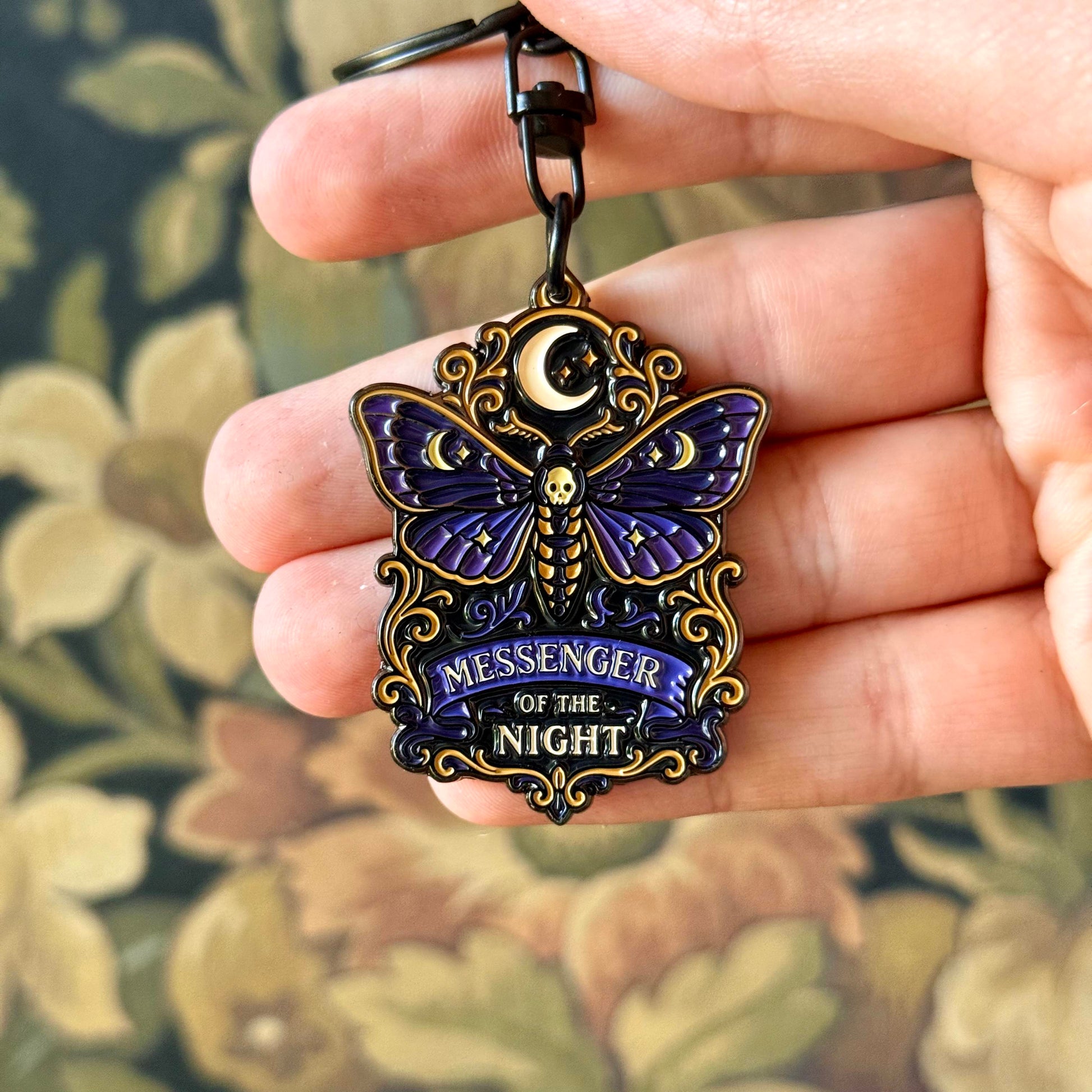 Moth keychain with 'Messenger of the Night' text held in a hand against a floral background
