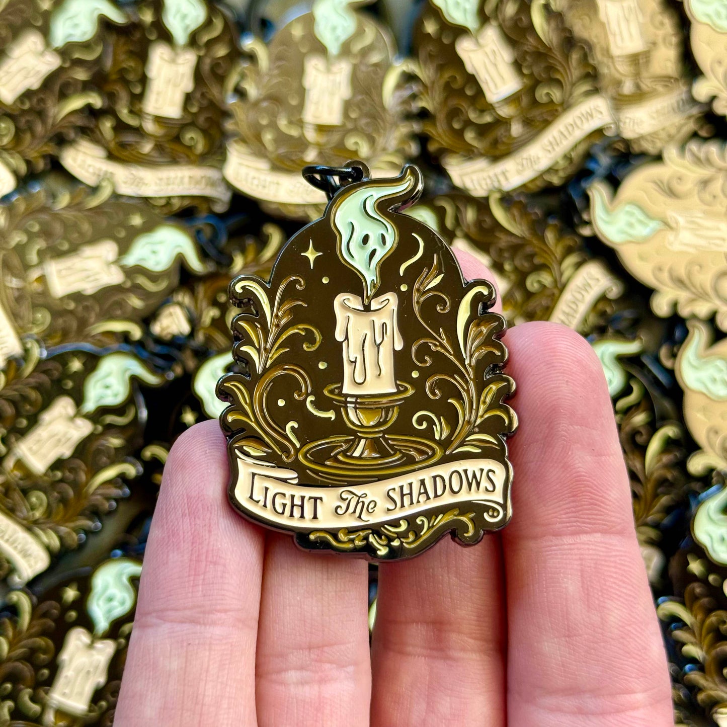 Decorative keychain with 'Light the Shadows' text held in a hand, with more keychains in the background.