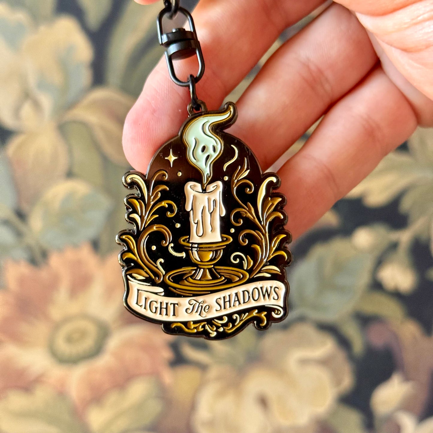 Decorative keychain with candle design and 'Light the Shadows' text held by a hand against a floral background.
