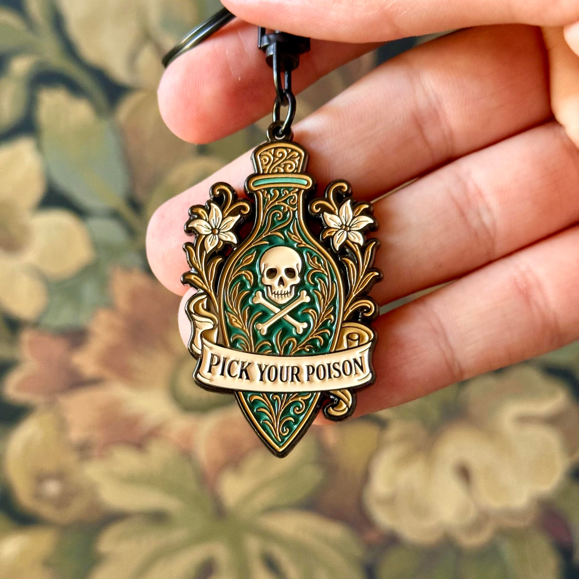 Keychain with skull design and 'Pick Your Poison' text held in a hand against a floral background