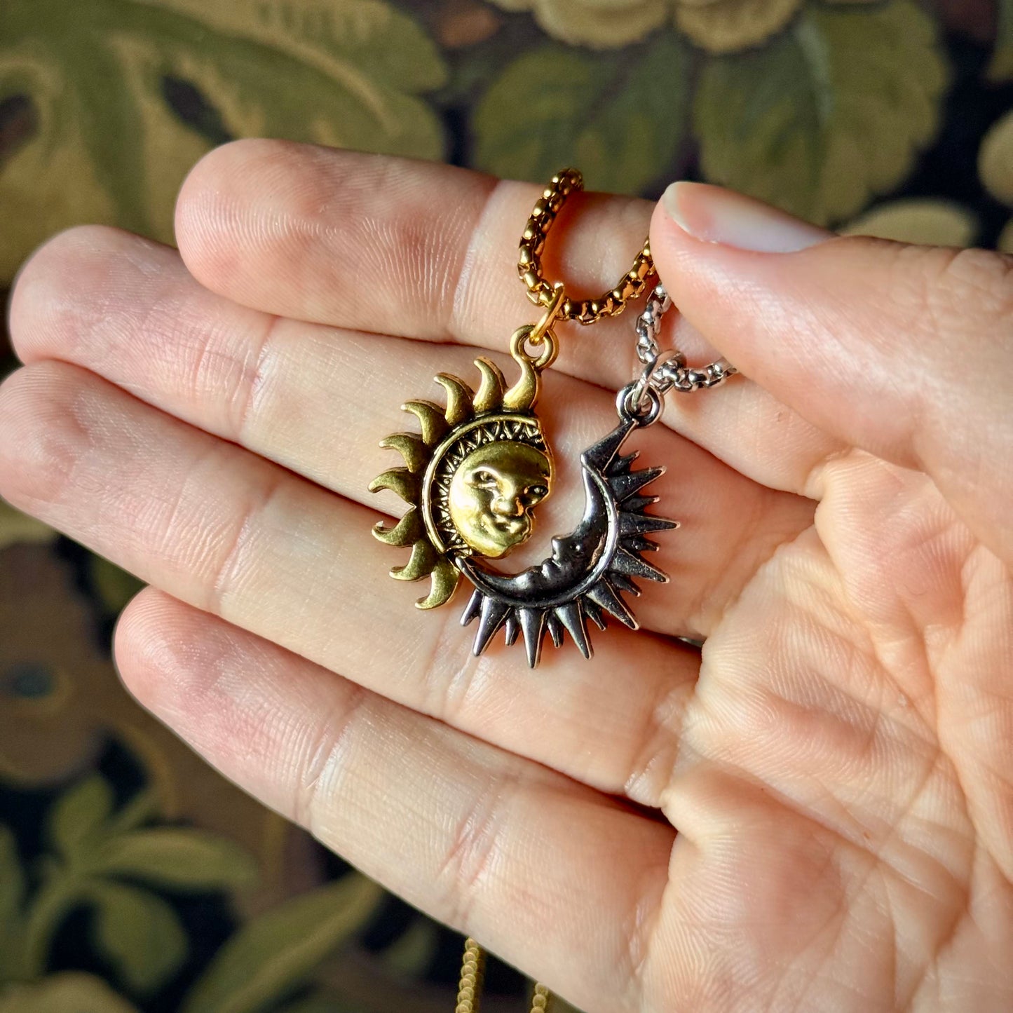 Two sun and moon pendants on a chain held in a hand against a floral background.