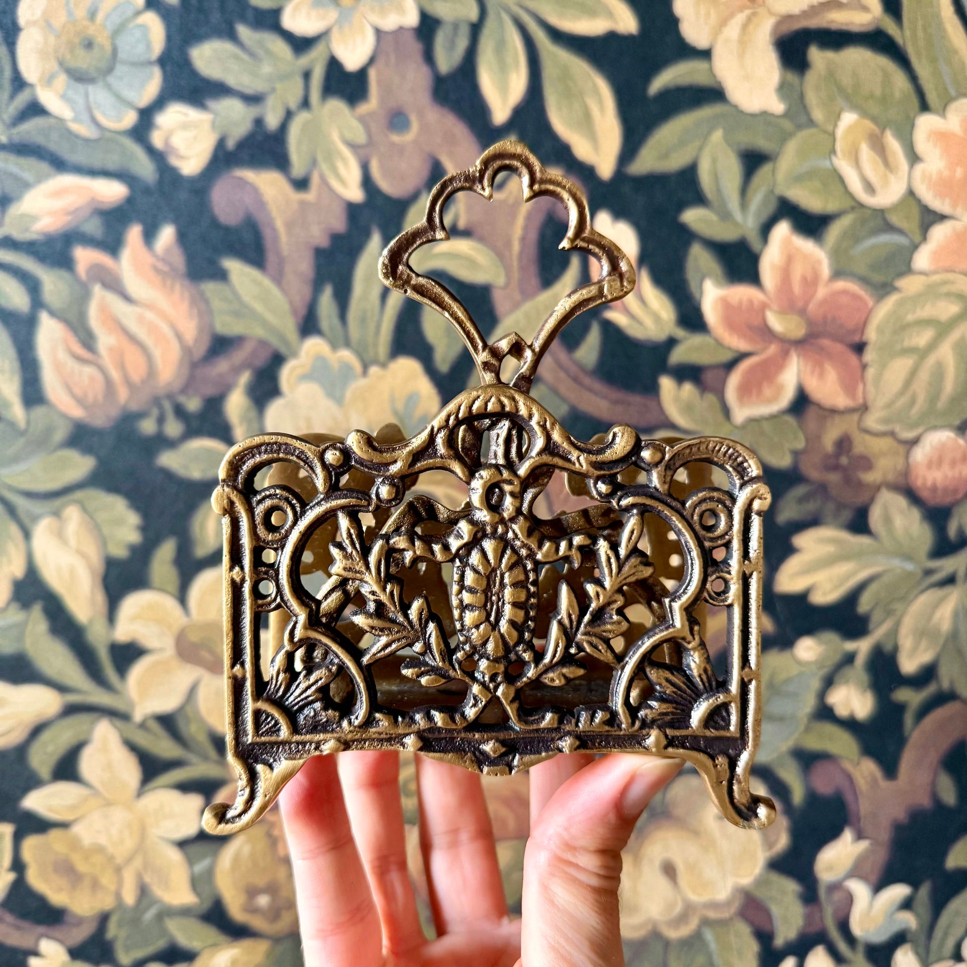 A detailed close-up of the Antique Brass Napkin and Letter Holder, showing its ornate curves and aged brass patina on a lace tablecloth.