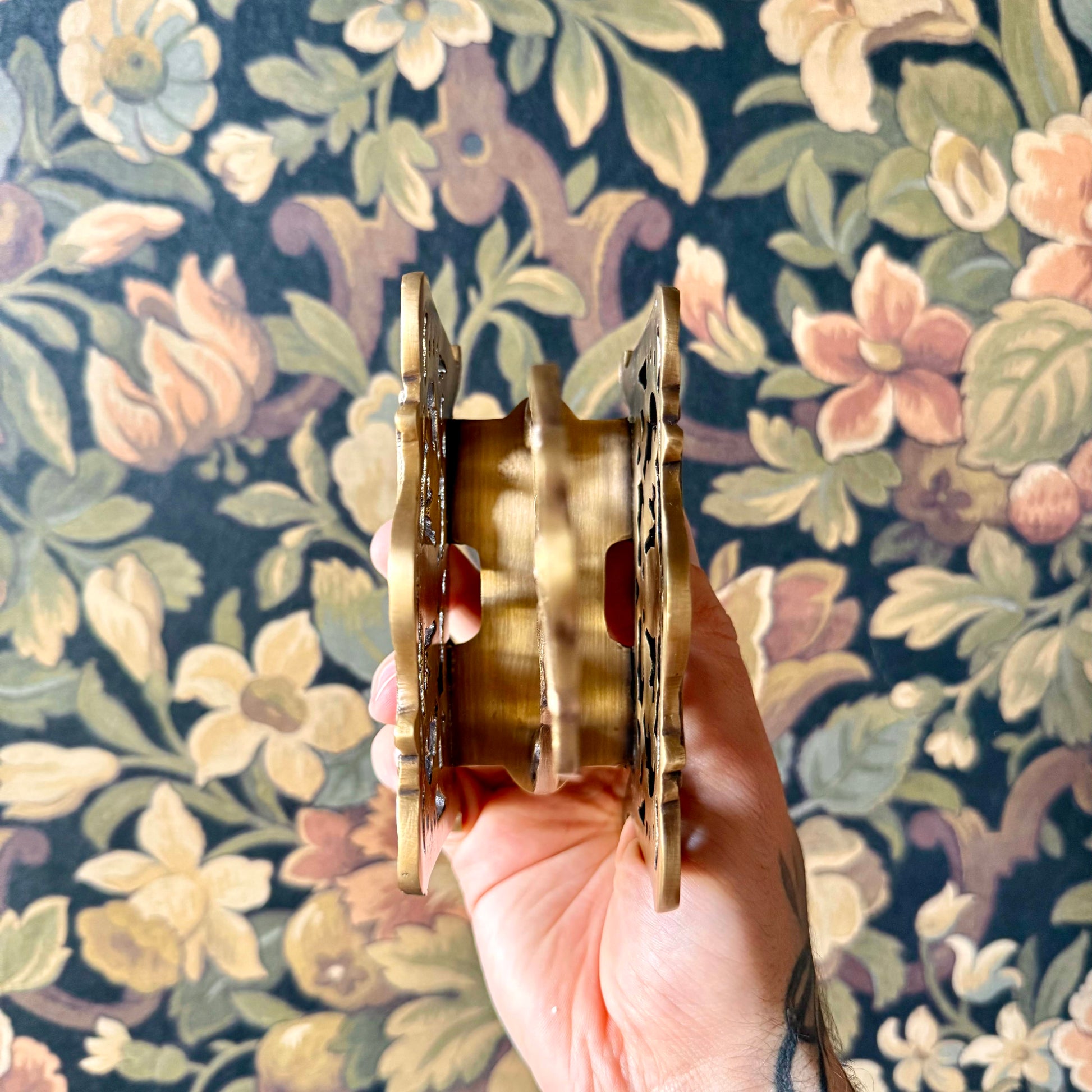 A detailed close-up of the Antique Brass Napkin and Letter Holder, showing its ornate curves and aged brass patina on a lace tablecloth.