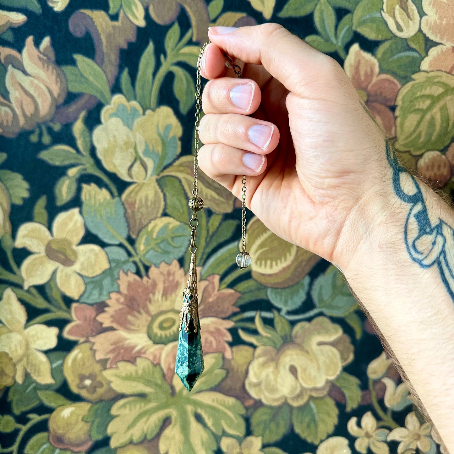 Victorian-inspired malachite pendulum featuring detailed brass accents. The malachite stone, with its rich green hues and natural banding, is suspended from a brass chain, ideal for spiritual practices or vintage-style decor.