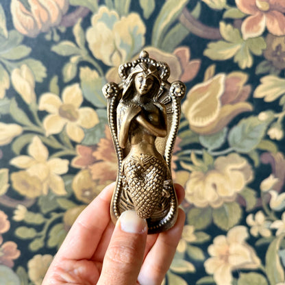 Antiqued brass coat hanger featuring a detailed mermaid figure with flowing hair and etched design, mounted on a neutral background. The vintage-style hook is solid and sculptural, measuring 5 inches in length.
