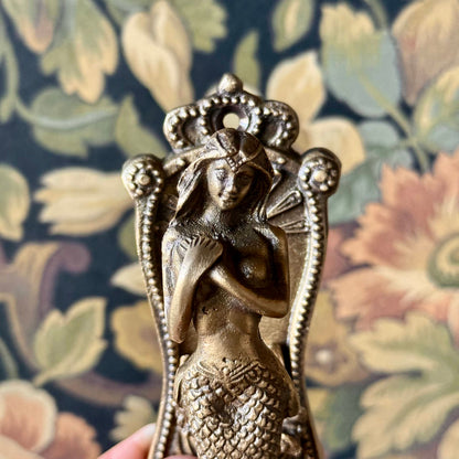 Antiqued brass coat hanger featuring a detailed mermaid figure with flowing hair and etched design, mounted on a neutral background. The vintage-style hook is solid and sculptural, measuring 5 inches in length.