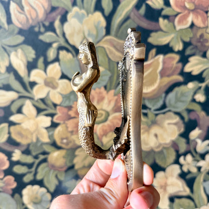 Antiqued brass coat hanger featuring a detailed mermaid figure with flowing hair and etched design, mounted on a neutral background. The vintage-style hook is solid and sculptural, measuring 5 inches in length.