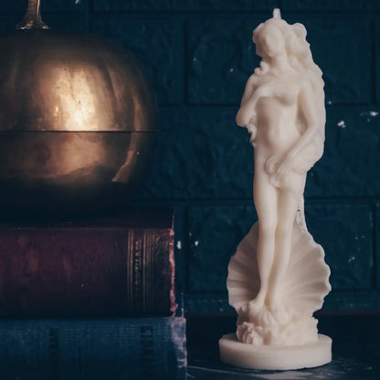 An off-white wax candle of classical female figure Venus standing on a scallop shell, displayed next to a brass pumpkin and old books against a dark textured background.