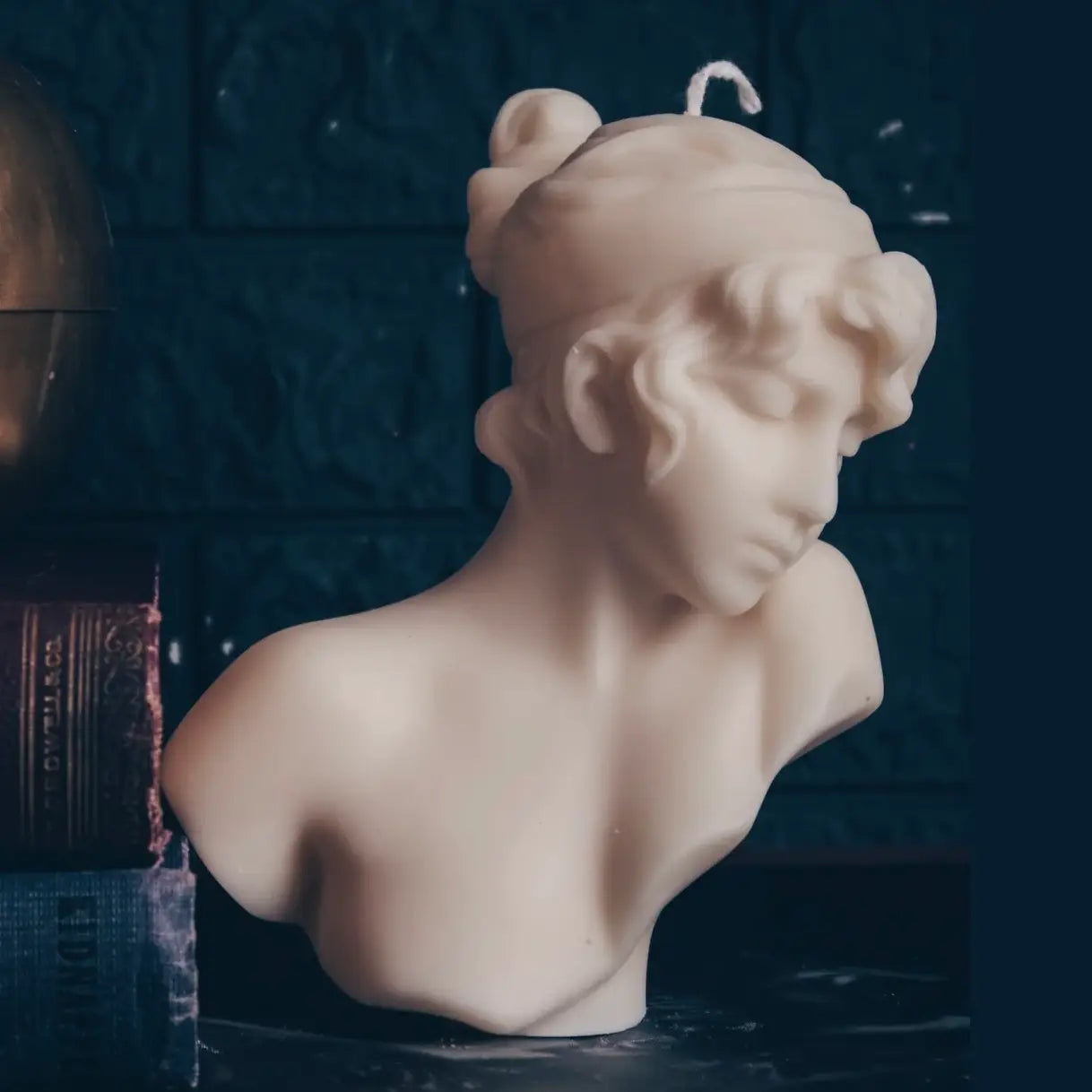 A beige sculptural bust candle shaped like a classical female bust with curly hair, photographed against a dark blue textured background with decorative books on the left.