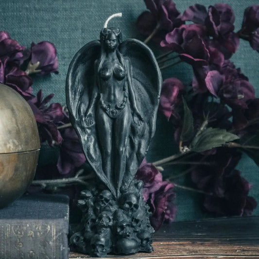 A black gothic winged female figure candle with a wick on top, standing on a skull-like base. The candle is styled on a wooden surface with dark purple flowers, a brass globe, and a textured teal background.