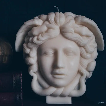 A sculptural candle shaped like the Medusa head, made of off-white wax with detailed curls and a single wick at the top, photographed against a very dark background.