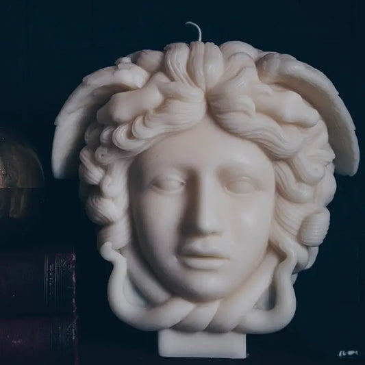 A sculptural candle shaped like the Medusa head, made of off-white wax with detailed curls and a single wick at the top, photographed against a very dark background.