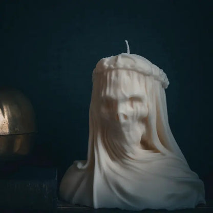A sculptural white candle shaped like a draped female figure or ghostly bust, with a single wick at the top, photographed against a very dark blue background.