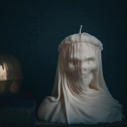 A sculptural white candle shaped like a draped ghostly or skeletal figure with a crown, photographed against a very dark blue background. A metallic object is partially visible on the left edge.