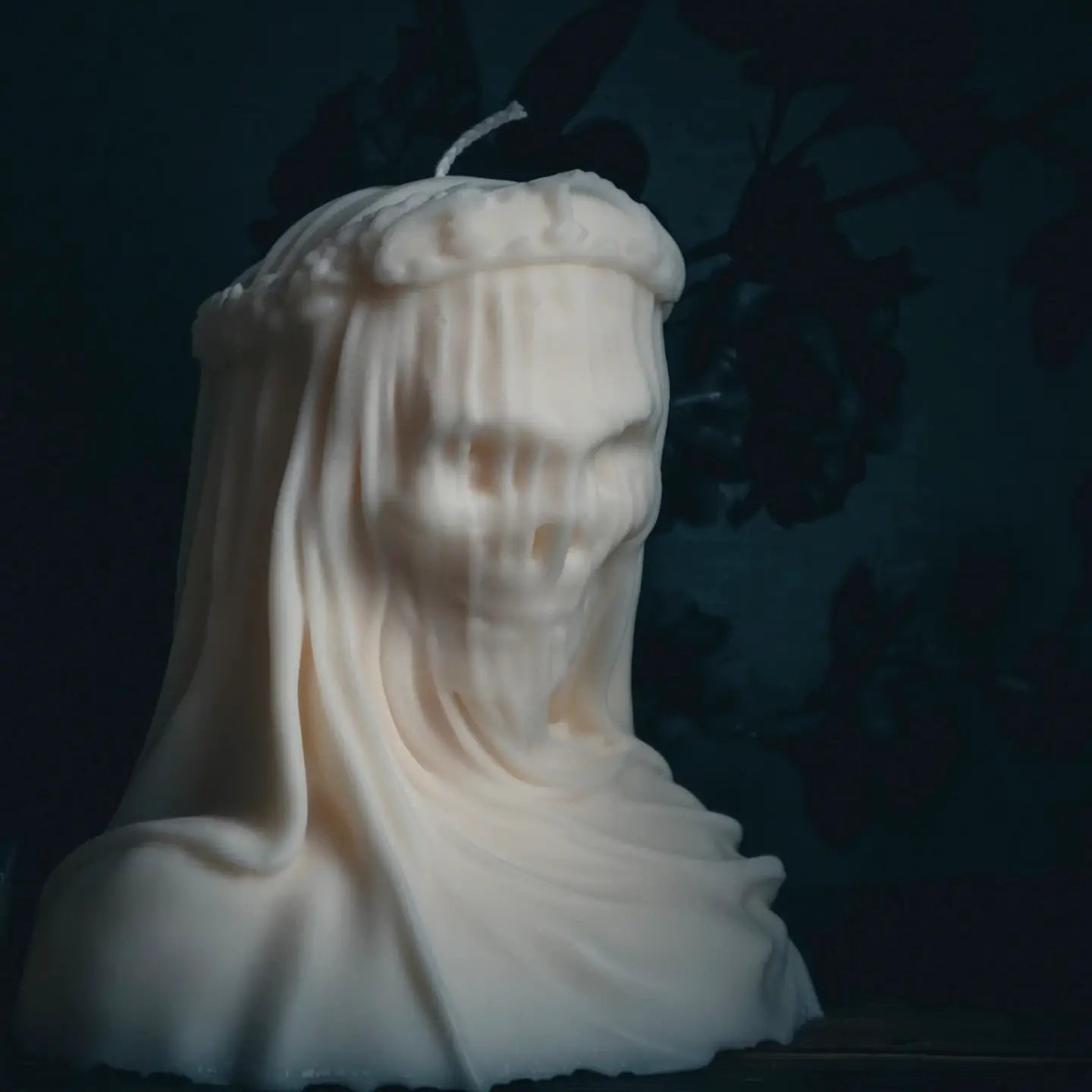 A sculptural white candle shaped like a draped ghost or hooded figure, photographed against a very dark blue background with faint shadowy floral shapes.