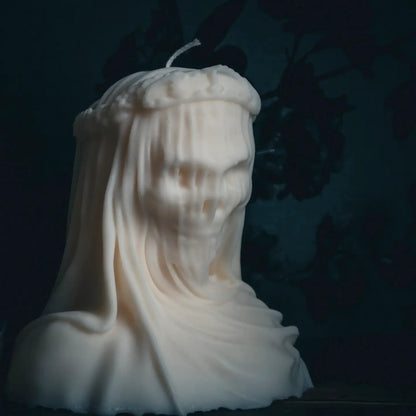 A sculptural white candle shaped like a draped ghost or hooded figure, photographed against a very dark blue background with faint shadowy floral shapes.