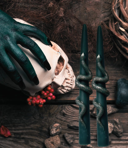 Two dark teal taper candles with metallic silver snakes wrapped around their sides, displayed on a rustic dark wood surface next to a skull and a hand wearing a matching dark teal glove.