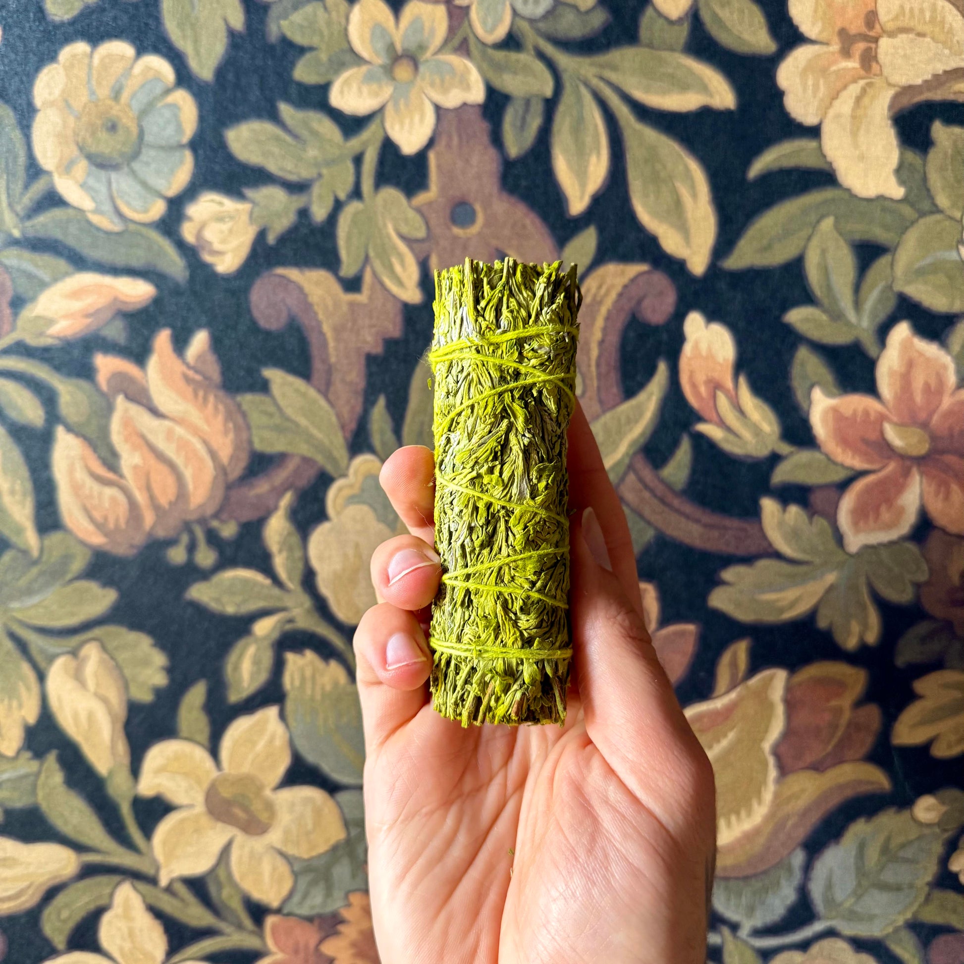A close-up view of a handcrafted Patchouli Resin Blue Sage Smudge Stick, showcasing its tightly bound, organic blue sage and patchouli resin details against a rustic wallpaper surface.