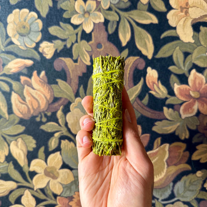 A close-up view of a handcrafted Patchouli Resin Blue Sage Smudge Stick, showcasing its tightly bound, organic blue sage and patchouli resin details against a rustic wallpaper surface.