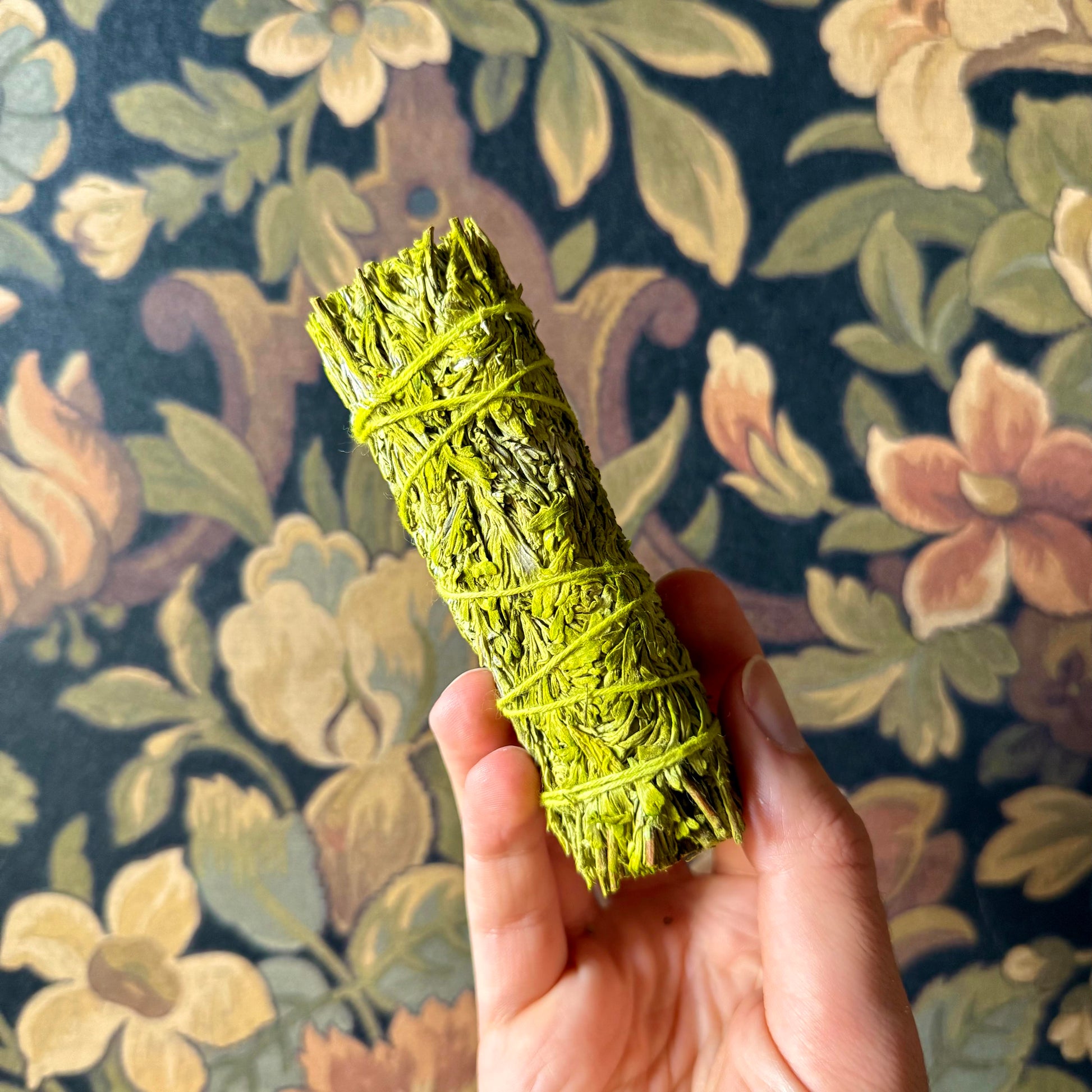 A close-up view of a handcrafted Patchouli Resin Blue Sage Smudge Stick, showcasing its tightly bound, organic blue sage and patchouli resin details against a rustic wallpaper surface.