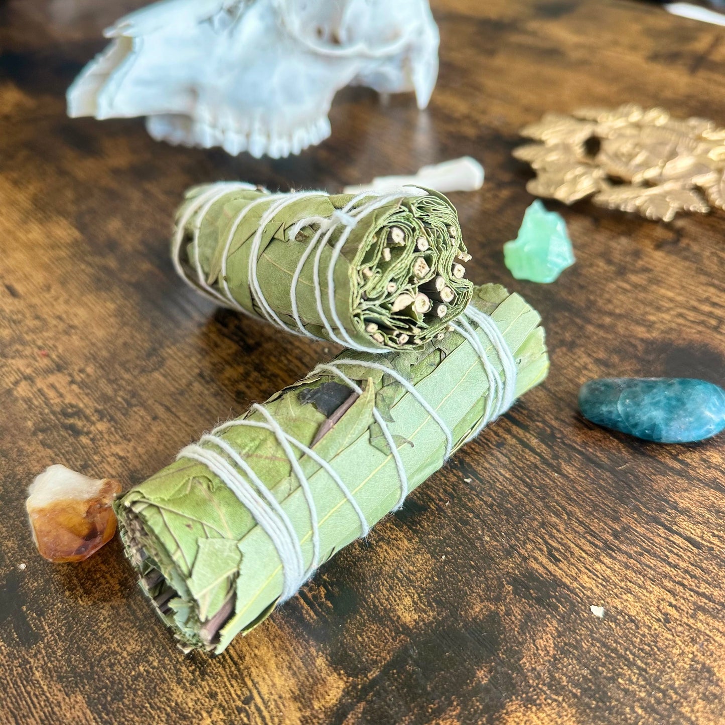 A close-up image of a eucalyptus smudge stick. The bundle features dried eucalyptus leaves tightly wrapped together with natural twine, forming a cylindrical shape. The leaves are a muted green color, with some light brown tones where they have dried. The smudge stick gives off an earthy and cleansing aura, perfect for rituals or purification practices. The texture of the leaves and twine is clearly visible, adding a natural, rustic feel to the image.