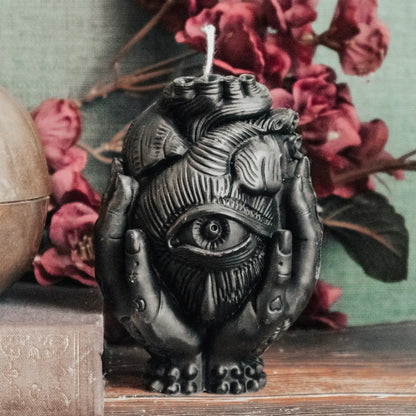 A black wax candle sculpted as a anatomical heart with an all-seeing eye on the front, cradled by two hands, displayed on a wooden surface with pink flowers and a muted green background.