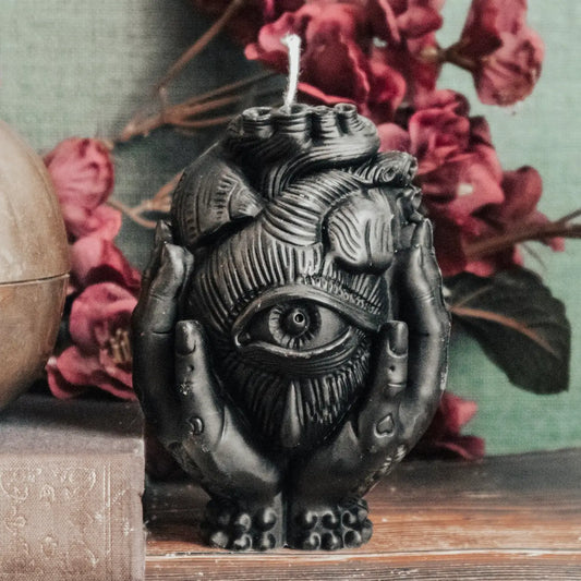 A black wax candle sculpted as a anatomical heart with an all-seeing eye on the front, cradled by two hands, displayed on a wooden surface with pink flowers and a muted green background.