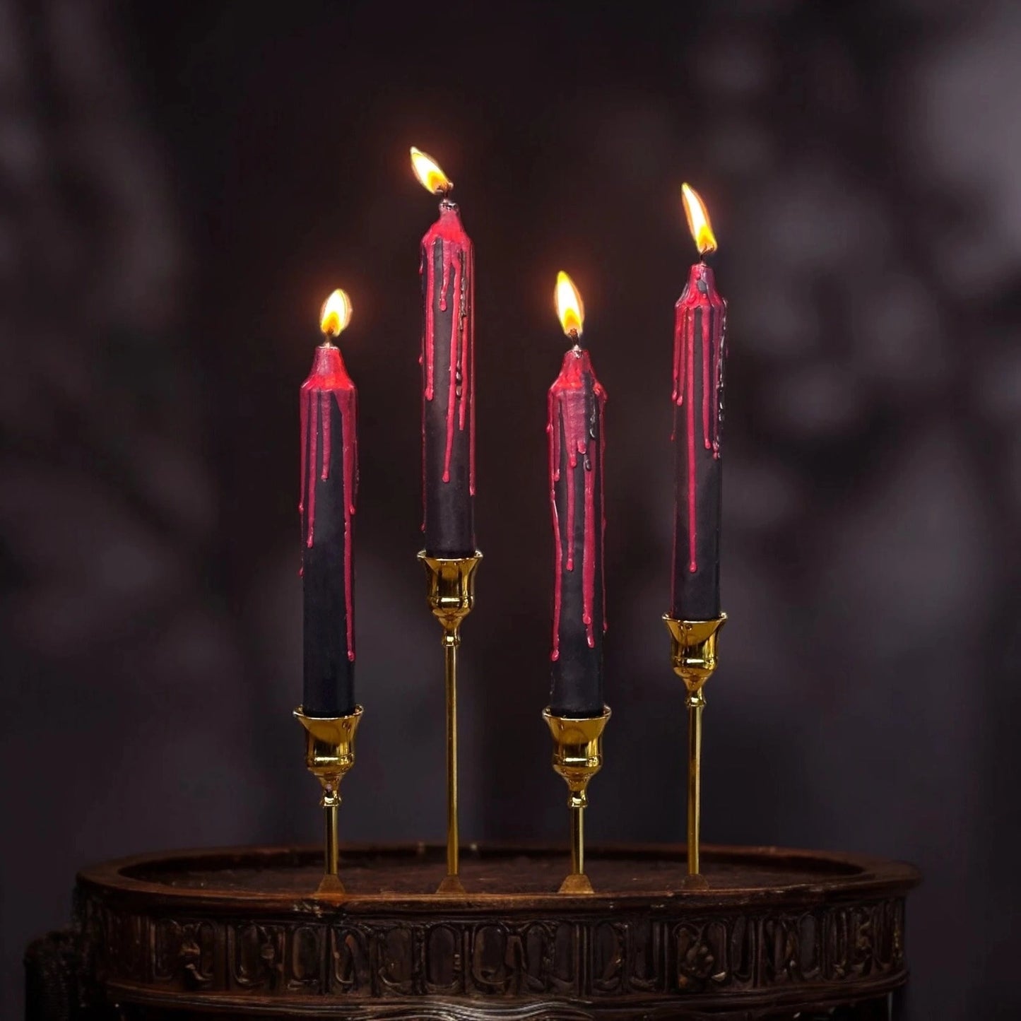 Four black taper candles with dripping red wax, lit and displayed in gold candlesticks on a dark wooden surface against a dark blurred background.