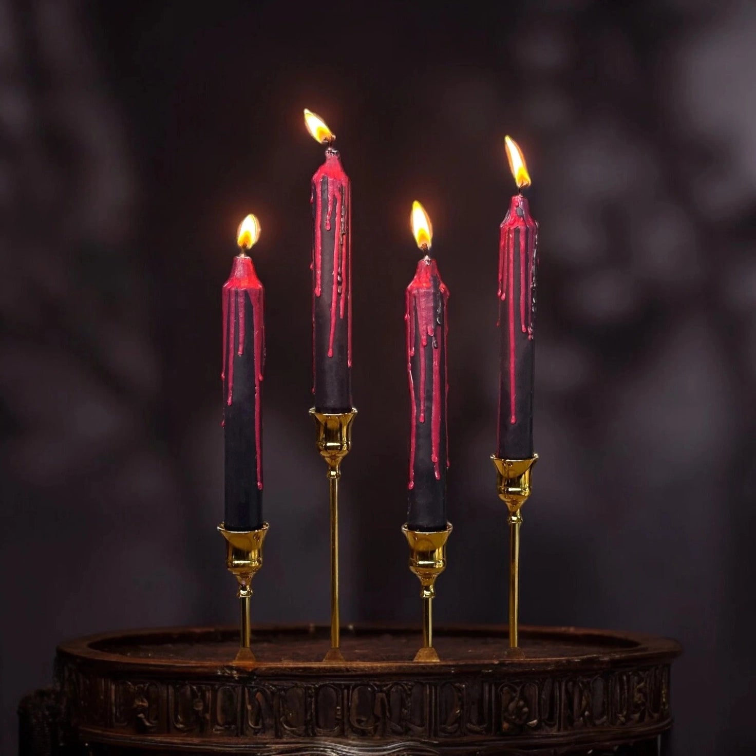 Four black taper candles with dripping red wax, lit and displayed in gold candlesticks on a dark wooden surface against a dark blurred background.